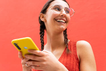 Von unten fröhliche junge Frau in Zöpfen Frisur Surfen auf Smartphone stehend Blick weg auf rotem Hintergrund in der Straße - ADSF28805