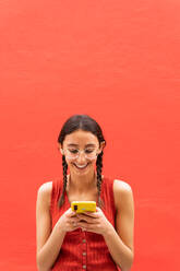 Fröhliche junge Frau in Zöpfen Frisur Surfen auf Smartphone stehend auf rotem Hintergrund in der Straße - ADSF28806