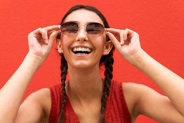 Erfreute junge Frau mit Zöpfen, die eine trendige Sonnenbrille vor rotem Hintergrund in einer Straße in der Stadt aufsetzen - ADSF28809