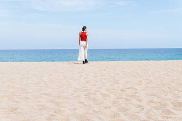 Rückenansicht einer Frau in Sommerkleidung, die an einem sonnigen Tag am Sandstrand auf das ruhige blaue Meer zugeht - ADSF28812