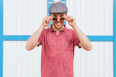 Junger bärtiger Mann in lässigem rotem Polohemd und Mütze, der über eine Sonnenbrille in die Kamera schaut und freundlich lächelt, während er im Sommer auf der Straße an der Wand steht - ADSF28822