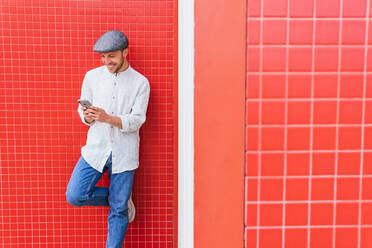 Zufriedener junger bärtiger Hipster in lässigem Poloshirt und Mütze, der im Sonnenlicht an einer roten Wand steht und auf seinem Handy surft - ADSF28823