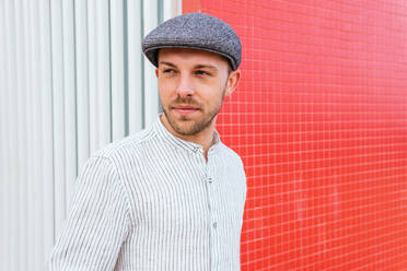 Hübscher junger bärtiger Mann in trendigem Freizeithemd, Jeans und Mütze, der in der Nähe einer rot-weißen Wand steht und mit einem Lächeln wegschaut - ADSF28824