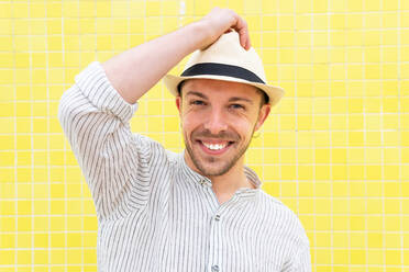Glücklicher junger bärtiger Hipster in trendigem Outfit und Hut, der lächelnd in die Kamera schaut, während er allein an einer gelben Wand im Sommer chillt - ADSF28827