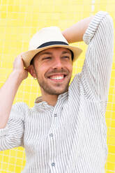 Glücklicher junger bärtiger Hipster in trendigem Outfit und Hut, der die Hände hinter dem Kopf hält und lächelt, während er allein an einer gelben Wand im Sommer chillt - ADSF28828