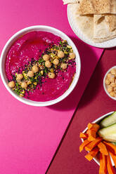 Top view of appetizing beetroot hummus garnished with chickpea served on two colored background with bread and fresh carrot and cucumber sticks - ADSF28830