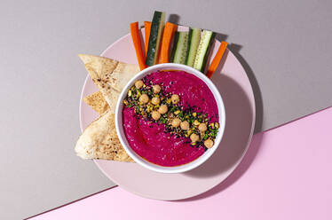 Top view of appetizing beetroot hummus garnished with chickpea served on two colored background with bread and fresh carrot and cucumber sticks - ADSF28832