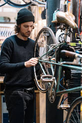 Seitenansicht eines seriösen männlichen Mechanikers, der ein Fahrradrad repariert, während er in einer Werkstatt arbeitet - ADSF28837