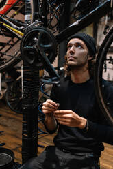Beschäftigter männlicher Meister, der ein modernes Fahrrad repariert, während er in einer schäbigen Werkstatt arbeitet und nach oben schaut - ADSF28839