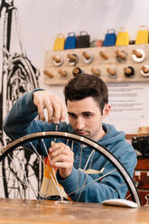 Seriöser männlicher Mechaniker, der an einer Werkbank sitzend eine Radspeiche repariert, in einer Fahrradwerkstatt - ADSF28843