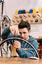 Serious male mechanic fixing wheel spoke while sitting at workbench in bike service - ADSF28843