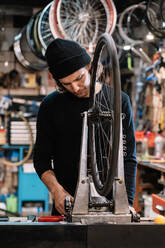 Young male master examining tire on bike wheel while working in professional repair service workshop - ADSF28844