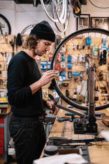 Seitenansicht eines jungen männlichen Meisters, der den Reifen eines Fahrrads bei der Arbeit in einer professionellen Reparaturwerkstatt untersucht - ADSF28847