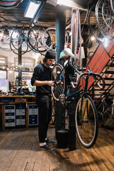 Junger männlicher Facharbeiter, der ein Detail am Rad eines Fahrrads repariert, das in der Werkstatt während der Wartungsarbeiten hängt - ADSF28848