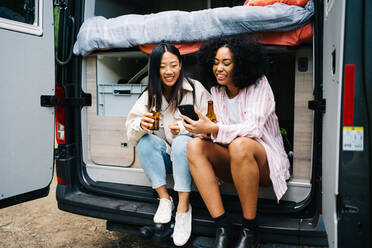 Multirassische junge Frau mit Bier, die auf ihrem Smartphone surft, während sie in einem Van auf einer Autoreise sitzt - ADSF28855