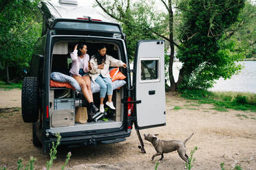 Reisende junge multirassische Freundinnen, die Bier trinken und dem Hund Befehle geben, während sie Spaß haben und sich gemeinsam in einem Wohnmobil ausruhen, das in der Nähe eines Flusses auf dem Lande geparkt ist - ADSF28858