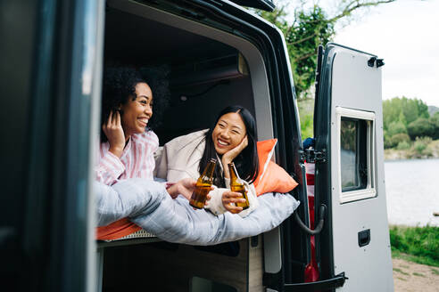 Fröhliche junge gemischtrassige Frauen trinken Bier, während sie im Wohnmobil während einer Sommerreise zusammen chillen - ADSF28859