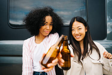 Happy young multiethnic female friends clinking bottles of beer while having fun and enjoying summer holidays together near camper van parked in nature - ADSF28868
