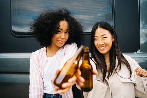 Glückliche junge multiethnische Freundinnen stoßen mit Bierflaschen an, während sie Spaß haben und die Sommerferien zusammen in der Nähe eines in der Natur geparkten Wohnmobils genießen - ADSF28868