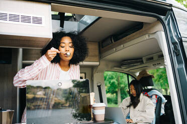 Junge schwarze Frau bereitet Kaffee zu, während sie ein Video auf dem Laptop im Wohnmobil mit einer asiatischen Freundin während einer Sommerreise in der Natur ansieht - ADSF28873