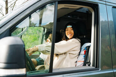 Durch das Fenster Seitenansicht einer fröhlichen jungen asiatischen Frau, die auf dem Fahrersitz eines Wohnmobils sitzt und in die Kamera schaut, während sie mit ihrer Freundin durch die Natur reist - ADSF28876