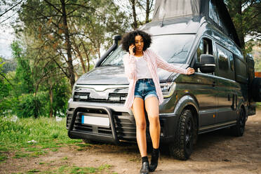 Serious young African American female talking on smartphone and gesticulating while leaning against camper car parked in forest in summer day - ADSF28880