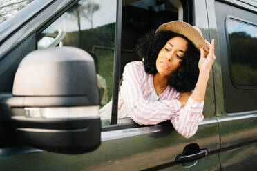 Positive young African American female in summer outfit and hat leaning out window of camper van and looking at side mirror while enjoying road trip - ADSF28882