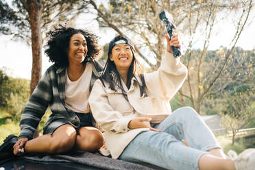 Low angle of cheerful young multiracial girlfriends sitting on roof of camper van and taking pictures while spending summer holidays together in countryside - ADSF28885