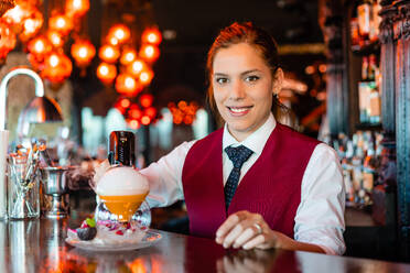 Geschickte junge Barkeeperin beim Garnieren von Cocktails am Bartresen mit einer Rauchpistole für Geschmacksverstärker - ADSF28890