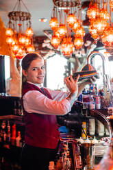 Seitenansicht einer weiblichen Barkeeperin in Uniform, die einen Cocktail in einem Metallshaker schüttelt, während sie am Tresen eines Pubs steht und in die Kamera schaut - ADSF28898