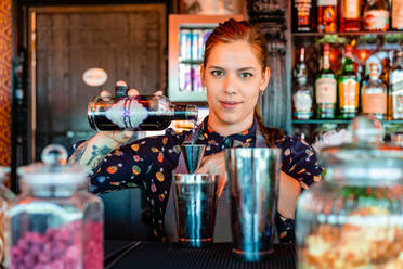 Zufriedene weibliche Barkeeperin, die Alkohol in einen Metallshaker gießt, während sie einen erfrischenden Cocktail am Tresen einer Bar zubereitet - ADSF28906