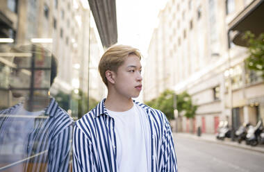 Stylish Asian male model standing near building and reflecting in glass mirrored wall - ADSF28923