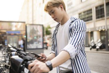 Focused ethnic male using sharing app on smartphone and renting bike parked on city street - ADSF28925