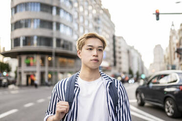 Trendy Asian male model with blond hair and backpack standing in city street and looking away - ADSF28927