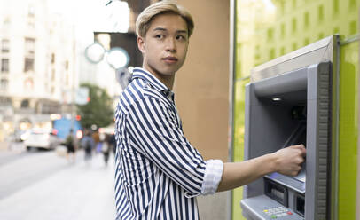 Seitenansicht eines ethnischen Mannes mit blondem Haar, der Geld an einem Geldautomaten in der Stadt abhebt - ADSF28929