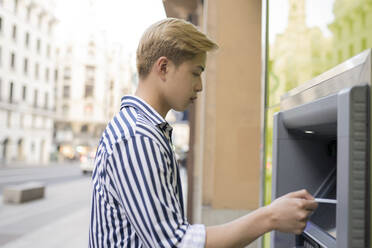 Side view of ethnic male with blond hair withdrawing money on ATM in city - ADSF28931