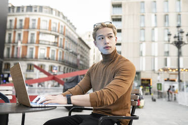 Nachdenklicher asiatischer männlicher Freiberufler, der mit einem Laptop an einem Tisch in einem Straßencafé sitzt und aus der Ferne an einem Startup arbeitet, während er wegschaut - ADSF28937