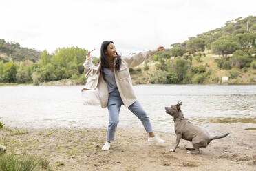 Ganzkörper asiatische Frau in Freizeitkleidung wirft Stock beim Spielen mit gehorsamen Hund am Ufer des Flusses in der Natur - ADSF28943