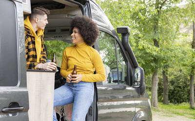 Happy black woman with beer smiling looking at each other standing in van with boyfriend during road trip in countryside - ADSF28949