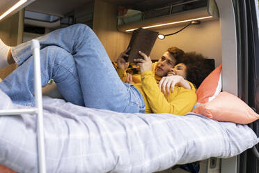 Unterschiedlicher junger Mann und Frau, die sich umarmen und ein interessantes Buch lesen, während sie auf dem Bett im Van während einer Autoreise chillen - ADSF28955
