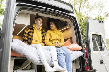 Happy black woman smiling and holding hands with boyfriend while sitting on bed inside van on weekend day in nature - ADSF28956