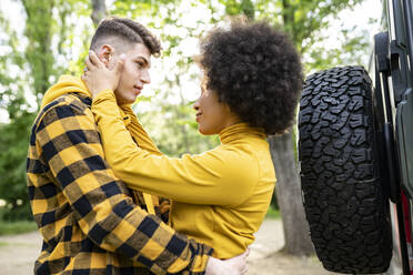 Niedriger Winkel eines jungen multiethnischen Mannes und einer Frau in Freizeitkleidung, die sich umarmen, während sie in der Nähe eines Autos an einem sonnigen Tag auf dem Lande stehen - ADSF28960