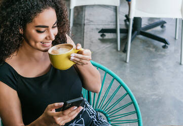 Von oben auf eine glückliche junge hispanische Frau, die auf ihrem Handy surft, während sie einen leckeren Cappuccino am Tisch eines Straßencafés genießt - ADSF28967