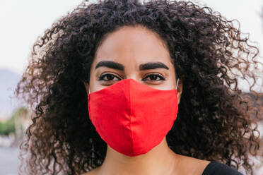 Young curly haired Hispanic female on red fabric protective mask for coronavirus prevention while spending summer day on street looking at camera - ADSF28968