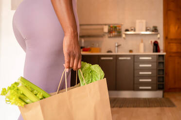 Side view of crop anonymous ethnic female with ripe green celery and lettuce in shopping bag standing in kitchen at home - ADSF28974