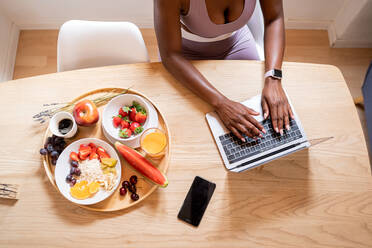 Oben auf dem Bild einer anonymen afroamerikanischen Frau, die auf einem Netbook tippt, während sie zu Hause am Tisch sitzt und ein gesundes Frühstück zu sich nimmt - ADSF28975