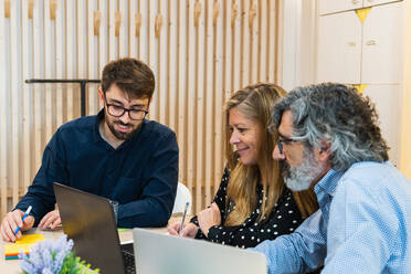 Delighted coworkers sitting at table during meeting in workplace and discussing project while looking at each other - ADSF28979