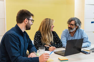 Delighted coworkers sitting at table during meeting in workplace and discussing project while looking at each other - ADSF28980