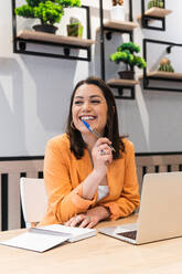 Smiling female freelancer sitting at table with laptop and notebook while thinking about startup project and looking away - ADSF28982