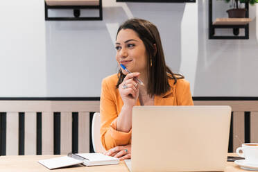 Smiling female freelancer sitting at table with laptop and notebook while thinking about startup project and looking away - ADSF28983